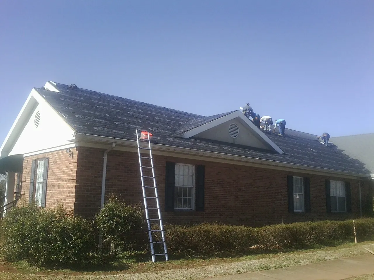 Crew tearing shingles off a brick ranch roof with a ladder set — Emergency Roof Repair work by our crew in Senoia