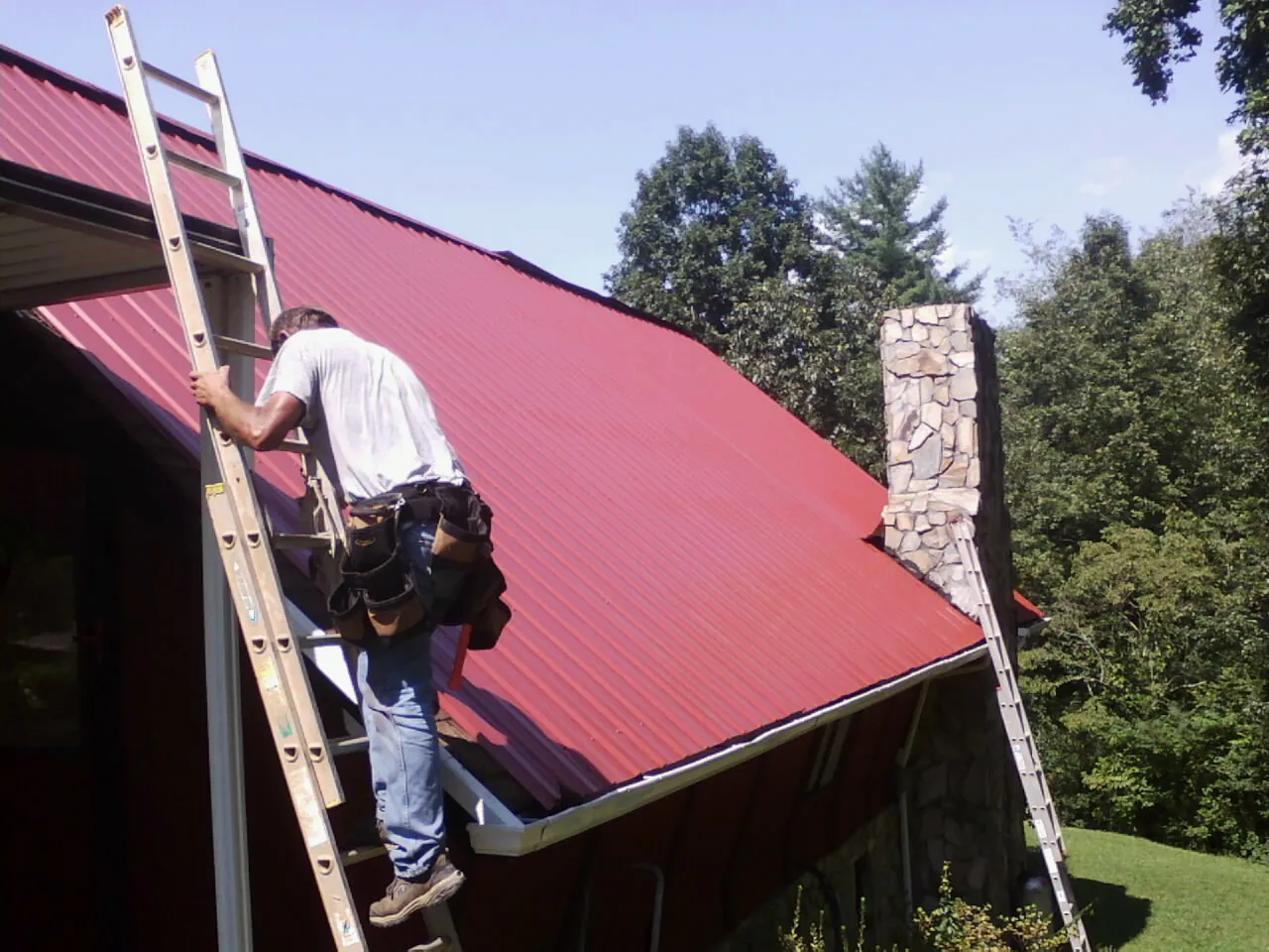 Roofer on a ladder beside a red metal roof and stone chimney — Soffit & Fascia Repair by Roofing Brothers in Senoia